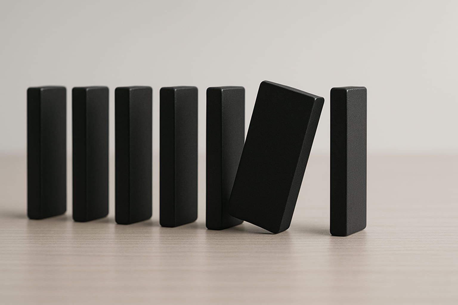 Seven black dominoes standing in a row on a pale wooden surface, the sixth tipped against its neighbour.